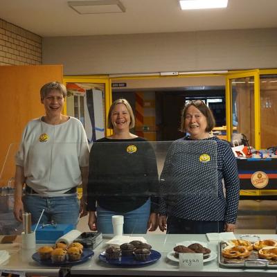 3 Frauen stehen hinter eine Theke Auf den Tischen der Theke stehen Kaffee, eine Kasse, Muffins und Brezeln zum Verkauf. 