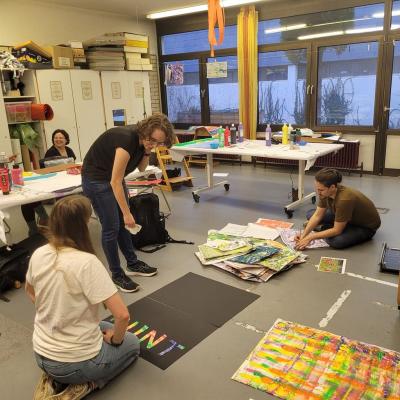 Im Kunstaktionsraum basteln Frau Fischer, Frau Blessing, Frau Huber und Herr Kling. Einige sitzen am Tisch, einige auf dem Boden. Überall sind bunte Plakate, Farben und Bastelmaterialien.