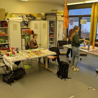 Im Kunstaktionsraum basteln Frau Seruga, Frau Bertz, Frau Blessing, Frau Fischer, Herr Michalek und Herr Kling. Überall sind bunte Plakate, Farben und Bastelmaterialien.