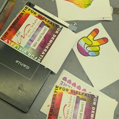 Auf dem Boden sieht man verschiedene bunt gestaltete Papiere mit Symbolen wie dem Peace-Symbol, die mit einer Schneidemaschine zurechtgeschnitten werden. Darauf steht zum Beispiel „Wir sind bunt!“, „Für Demokratie“ und „Für Vielfalt“.