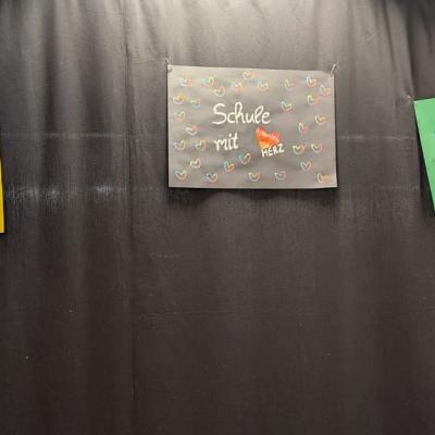 In der Aula an einem Vorhang hängen drei Plakate mit verschiedenen Botschaften. Darauf steht „Alle Haben Wert“ und „Schule mit Herz“. Rechts ist eine bunte Blüte zu sehen.
