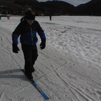 Josua hat einen Ski am rechten Fuß angeschnallt. Der linke Fuß ist ohne Ski und Josua nutzt ihn zum Anschieben wie auf einem Roller. So bewegt er sich über den Schnee. Im Hintergrund sieht man die Loipe, waldgrüne Berge und ein paar Leute.