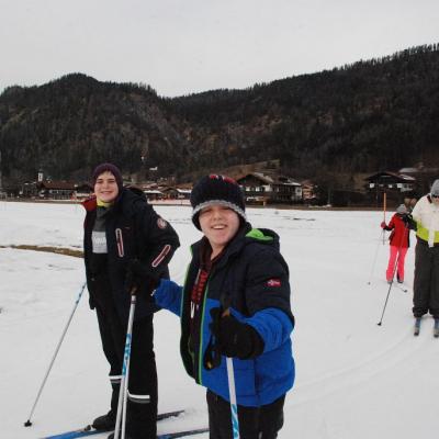 Auf der Loipe laufen Christopher, Josua, Julian und Lara. Die vorderen beiden lächeln in die Kamera. Seitlich steht ein Schild, das die Loipenrichtung vorgibt. Im Hintergrund sieht man bewaldete Hügel und Häuser.