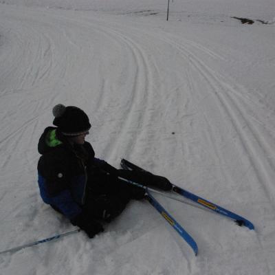 Josua sitzt seitlich auf der Loipe und schaut auf seine Ski. Er grinst.