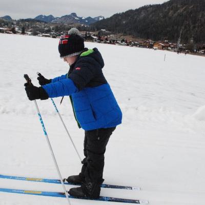 Josua steht in der Loipe und fährt vorwärts. Im Hintergrund sieht man eine Schneefläche. Dahinter sind Häuser und bewaldete Hügel.