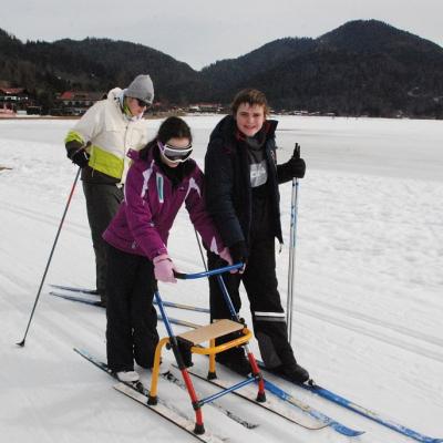 Am linken Bildrand steht Herr Kling und spricht mit den Schüler:innen. Auf den Loipen vor ihm stehen Christopher, Lara, Julian und Josua. Im Hintergrund sieht man das Schneefeld und bewaldete Hügel.