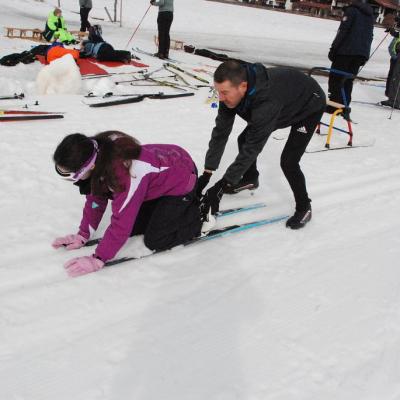 Marija kniet auf ihren Skiern und hat die Hände vorne auf den Skispitzen. Herr Schellinger von den Stuttgartern schiebt sie an den Fersen an.