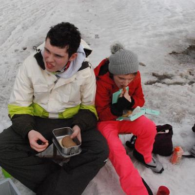 Julian und Lara sitzen auf einem Schlitten im Schnee und vespern.