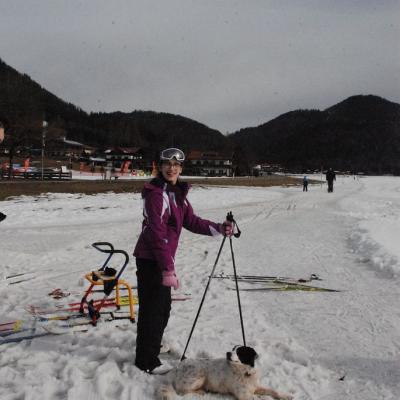 Marija steht mit Skistöcken in der Hand im Schnee. Vor ihr liegt ein Hund im Schnee und schaut zu ihr hoch. Im Hintergrund sieht man bewaldete Hügel.