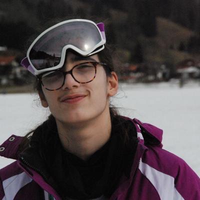 Marija lächelt in die Kamera. Ihre Skibrille hängt schief auf ihrer Stirn. Im Hintergrund sieht man unscharf die Landschaft.
