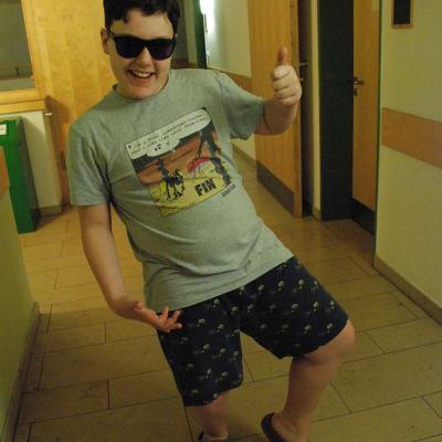 Josua trägt T-Shirt und kurze Hose im Flur und eine Sonnenbrille. Er lächelt und zeigt Daumen hoch.