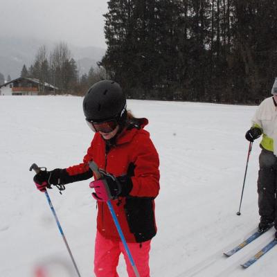 Lara und Julian laufen hintereinander auf der Loipe. Dahinter ist alles weiß von Schnee und man sieht den Waldrand. Der Himmel ist bewölkt.
