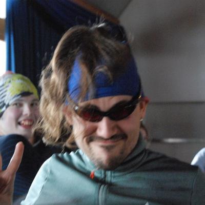 Herr Kling hat sein Halstuch um den Kopf gebunden, eine rote Sonnenbrille auf und seine langen Haare vorne ins Gesicht. Dazu macht er eine Grimasse.