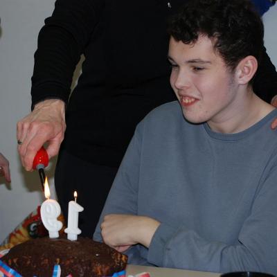 Julian sitzt vor einem Kuchen, auf dem Kerzen in Form der Zahl 19 stecken. Er lächelt freudig. Frau Gauch hat eine Hand auf seiner Schulter und zündet mit der anderen die Kerzen an.