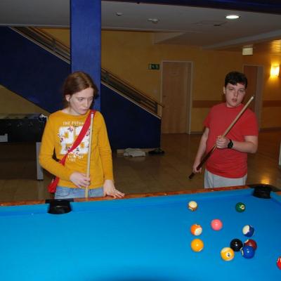 Lara und Josua stehen am Billardtisch und schauen auf die Kugeln. Beide halten einen Billard-Queue in der Hand. Im Hintergrund sieht man einen Tischkicker und eine Treppe.