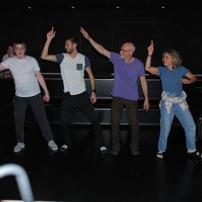 Christopher und einige Lehrer:innen von verschiedenen Schulen stehen im dunklen Disco-Raum und tanzen eine Choreographie. Alle halten den rechten Arm nach oben.