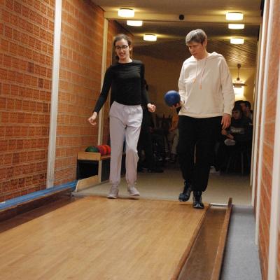Marija und Frau Gauch laufen nebeneinander auf die Kegelbahn zu. Frau Gauch trägt eine blaue Kugel. Sie erreichen gerade das Parkett der Kegelbahn.