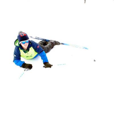 Josua liegt auf dem Bauch im Schnee und stützt sich mit den Armen ab. Er lächelt in die Kamera. Er hat seine Stöcke in den Händen und seine Langlaufskier an die Füßen.