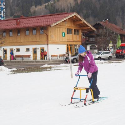 Marija läuft mit Langlaufskiern und ihrem Skilator auf der Loipe. Im Hintergrund sieht man eine Touristeninformation in einem großen Holzhaus. 