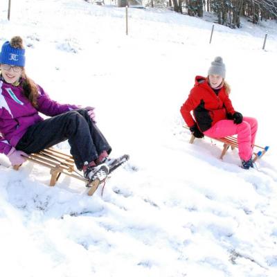 Marija (links) und Lara (rechts) sitzen am Hang jeweils auf einem eigenen Schlitten. Sie haben ihre Skiklamotten an und schauen in die Kamera. 