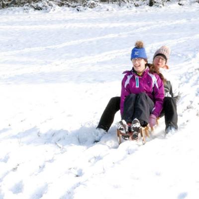 Marija und Frau Schremger fahren gemeinsam auf einem Schlitten schnell den verschneiten Berg hinunter. Sie scheinen große Freude zu haben. 