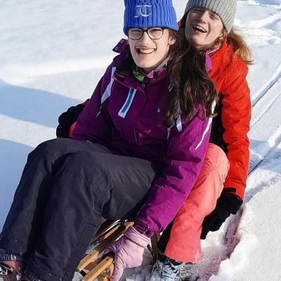 Marija und Lara sitzen gemeinsam auf einem Schlitten. Sie lachen in die Kamera. Rings herum ist Schnee zu sehen. Ihr Schlitten hat spuren im Schnee hinterlassen. 