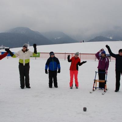 Sechs Personen stehen in einem Halbkreis und sind in Bewegung mit ihren Armen. Sie stehen neben der Loipe im Schnee und machen Aufwärmspiele. 