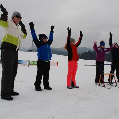 Sechs Personen stehen mit nach oben gestreckten Armen im Halbkreis. Der Boden ist mit Schnee bedeckt und man sieht Berge mit verschneitem Wald im Hintergrund. 