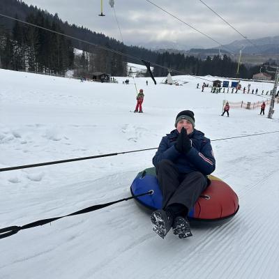 Christopher sitzt in einem Schlauchreifen, der von einem Snowtubinglift gezogen wird. Er hält die Hände zum Gebet gefaltet. Im Hintergrund ist ein Tellerlift.