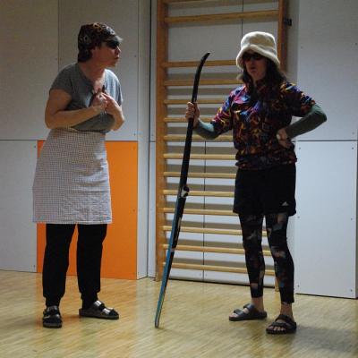 Lisel und Berta, stehen vor einer Sprossenwand. Lisel hält einen Langlaufski in der Hand. Sie unterhalten sich.