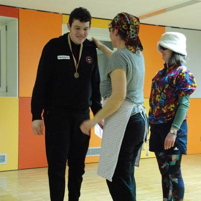 Berta legt die Hand auf Julians Schulter. Er hat eine Medaille um den Hals hängen. Lisel steht rechts daneben. 