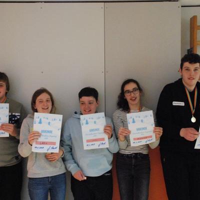 Alle fünf Schüler:innen der AHW halten ihre Medaillen und Urkunden in die Kamera. Sie stehen nebeneinander vor einer Wand. Alle fünf lachen. 