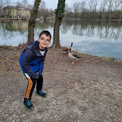 Ein Junge posiert lächelnd in die Kamera. Neben ihm stehen zwei Enten. Im Hintergrund ist ein See mit einem kleinen Schloss zu sehen.