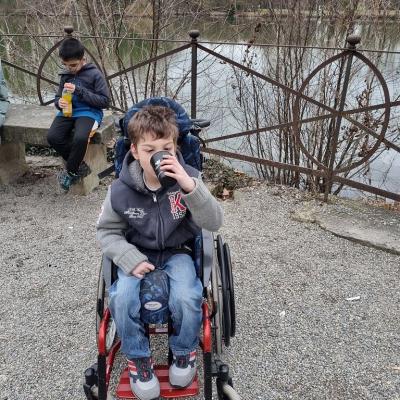 Ein Junge im Rollstuhl hält einen Becher in der Hand und trinkt. Hinter ihm sitzt ein Junge auf der Bank und trinkt ebenfalls