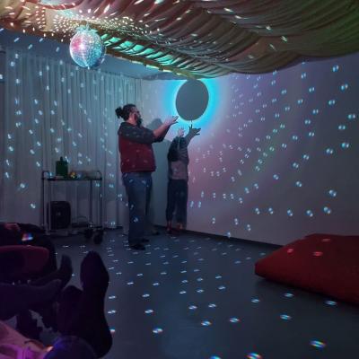 Zwei Erwachsene und ein Junge sitzen auf dem Boden. Sie fühlen in einer Kiste. Über der Kiste ist ein gelbes Tuch. Um die Personen sind ganz viele bunte Lichter. 