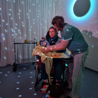 Ein Mädchen im Rollstuhl und eine Frau fühlen den Inhalt einer Kiste. Um sie herum sind ganz viele bunte Lichter