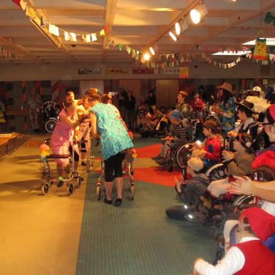 Fasching in der Schulaula, rechts viele verkleidete Kinder, mittig als Putzfrauen mit Rollatoren verkleidete Frauen bei einer Aufführung