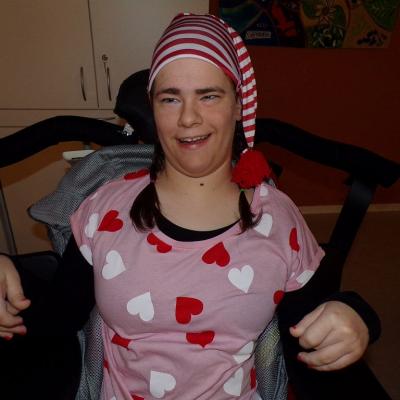 Ein Mädchen lächelt in die Kamera. Sie trägt ein Rosa Schlafanzug mit roten und weißen Herzen darauf. Sie hat geflochtene Zöpfe und trägt eine Rot/Weiß gestreifte Bommelmütze