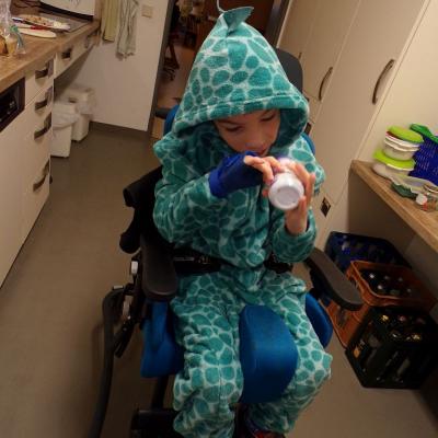 Ein Junge sitzt auf einem Stuhl. Er trägt ein grünes Dinosaurierkostüm. Er hat die Kaputze auf, auf dieser sind kleine Stacheln. Er spielt mit einer Flasche die er in der Hand hält. 