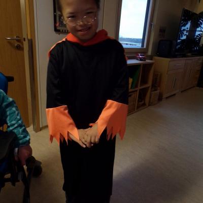 Ein Junge lächelt schüchtern in die Kamera. Er trägt ein Schwarzes Gewand mit Orangenen Ärmeln und Kragen. 