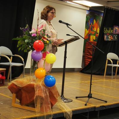 Die Schuldirektorin Frau Fißler steht auf der Bühne und hält eine Eröffnungsrede der Veranstaltung. Vor Ihr steht eine Vase mit einem Blumenstrauß, welche mit bunten Luftballons geschmückt ist.