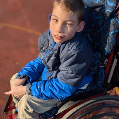 Ein Junge sitzt im Rollstuhl und lächelt in die Kamera. Die Sonne scheint und er steht auf einem Sportplatz