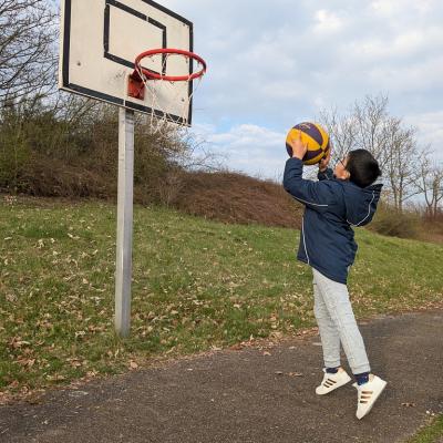 Es ist ein Junge von der Seite zu sehen. Er springt und wirft einen Ball. Vor ihm ist ein Basketballkorb