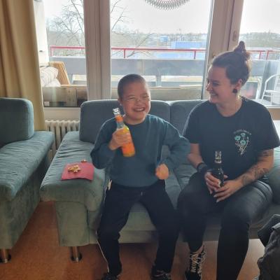 Ein Junge und eine Frau sitzen auf einer Couch. Beide lachen, der Junge schwingt freudig sein Arm. Beide halten Limonade in der Hand 