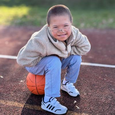 Ein Junge lächelt in die Kamera. Es ist schönes Wetter. Er sitzt auf einem Basketball auf einem Sportplatz.