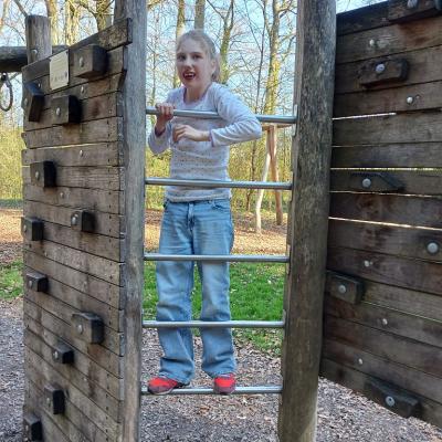 Schülerin mit blauer Jeanshose, grauen lang Arm Shirt steht auf einem Klettergerüst und lächelt mit ihren blinden Haaren in die Kamera. 