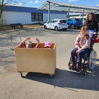 Zwei Schülerinnen sitzen im Holzwagen, man sieht nur zwei Köpfe , eine jubelt mit nach oben gestreckten Armen und die andere schaut über die Kante, rechts neben der Kiste ist eine Rollstuhlfahrerin und lacht darüber 