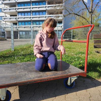 Schülerin sitzt auf dem Transportwagen und lacht. Im Hintergrund ist das Internat zu sehen