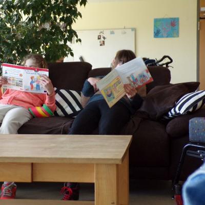 Zwei Schülerinen sitzen auf dem braunen Sofa auf der Grupppe, die Wand ist gelbgestrichen , es liegen 2 gestreifte Kissen auf dem Sofa in schwarz-weiß gestreift. 