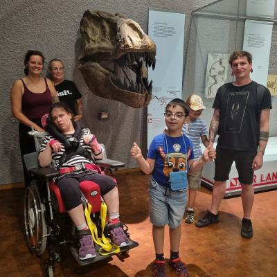 Ein Mädchen und zwei Jungen stehen mit einem Mann und zwei Frauen im Museum. Hinter ihnen ragt der Schädel eines Tyrannosaurus Rex aus der Wand.